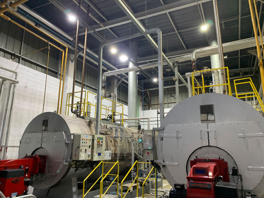 dpcex-sala-calderas Sala de una fábrica conde aparecen dos calderas de dimensiones grandes con conductos conectados a ellas.